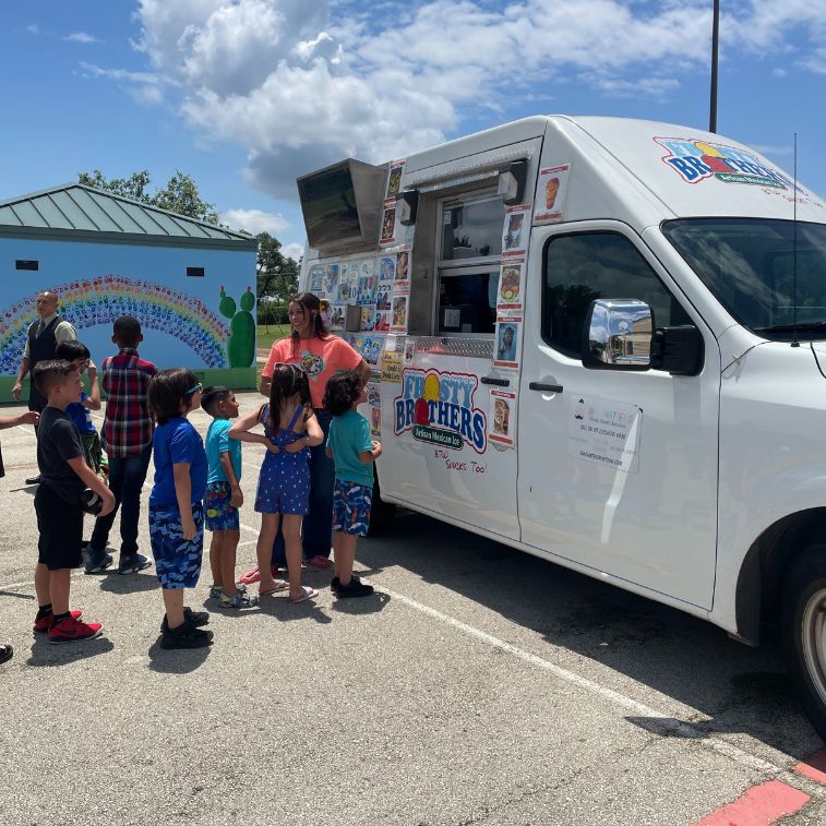 Children lined up at the Frosty Brothers Ice Cream Truck enjoying shaved ice and frozen treats during a school event in San Antonio, Texas.