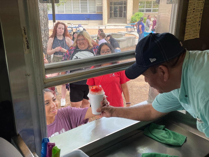 Carlos Sr. handing a fresh cherry shaved ice to a customer through the Frosty Brothers Ice Cream Truck service window at a catering event.